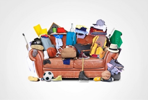 A cluttered assortment of household items and sports equipment piled atop a brown fabric sofa, with various clothing items, shoes, and a basketball visible among the mess. The clutter includes a yellow plastic container, a small chalkboard, a soccer ball, a badminton racket, and a baseball cap, with additional clothing such as jeans, shirts, and a hat scattered around. The background is plain and neutral, emphasizing the disorganized state of the items, which appear ready for rubbish removal. The scene suggests an urgent need for clearing and disposal, typical of a waste management service in the Highgate area, handled by Commercial Waste Removal Highgate, specialising in rubbish removal for domestic and commercial properties.