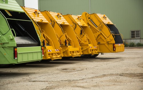 A row of four large commercial waste collection vehicles parked on a gravel surface in front of a green industrial warehouse. The first vehicle on the left is painted in a green color with a black loading mechanism, while the remaining three vehicles are bright yellow with black and silver hydraulic compactor elements. Each vehicle has a large, boxy body with visible hinges and locking mechanisms for the waste compartment, and their rear ends are designed for waste loading or compaction. The vehicles are arranged side by side, aligned along the paved area, with the warehouse wall featuring a small rectangular window and a vent near the right edge of the image. The scene is evenly lit, indicating daylight conditions, and presents a professional and functional environment typical of commercial waste management operations in Highgate or nearby London postal code areas, consistent with services offered by Commercial Waste Removal Highgate.