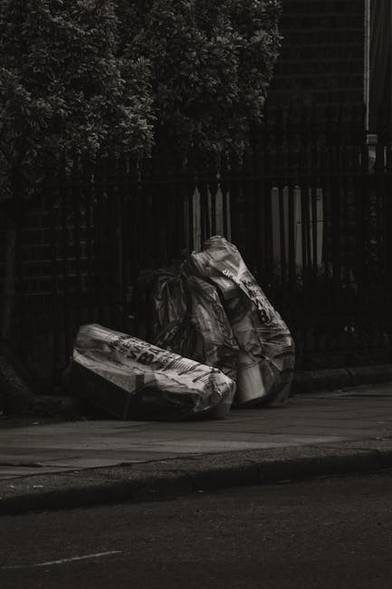 A stack of large garbage bags filled with waste material, positioned on the edge of a street pavement. The bags are made of black plastic with some transparent sections, revealing mixed debris inside.