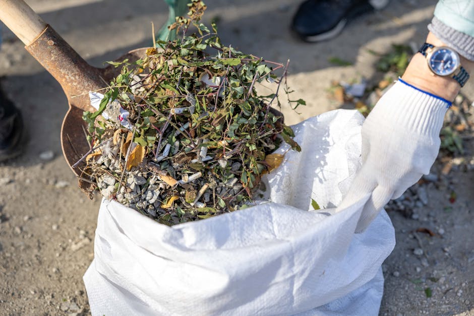 A small, light blue pickup truck parked on the side of a paved road, loaded with a large pile of mixed waste materials including cardboard boxes, black plastic bags, wooden planks, and other debris. T