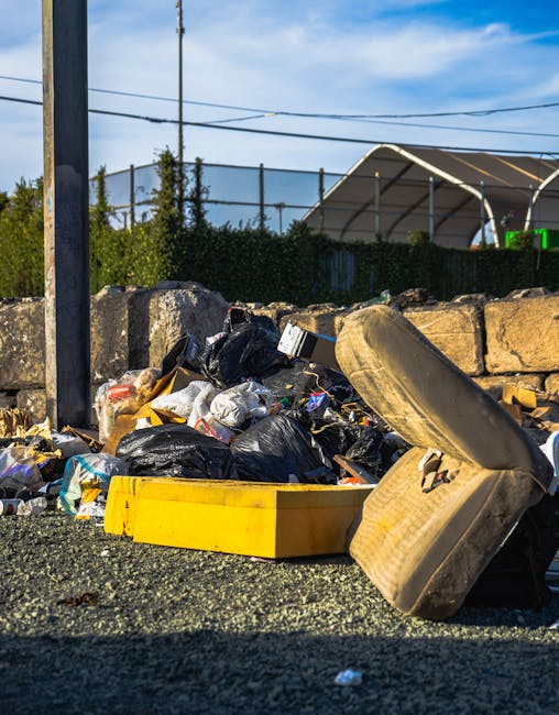 A group of black refuse bags filled with waste are stacked on a paved sidewalk near a black metal fence, with individual bags appearing crumpled and loosely packed. Behind the bags, there is a dark gr